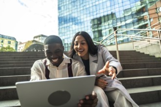 Diverse business colleagues sitting on steps, smiling and laughing while looking at a laptop