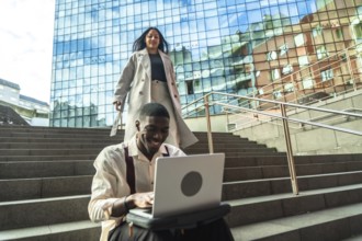 Young professional smiling at laptop on urban steps while a stylish woman walks down nearby by a