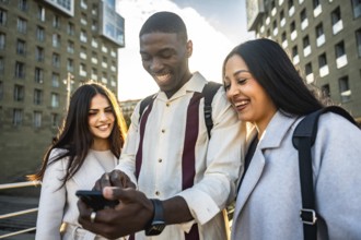 Diverse group of young friends laughing and sharing smartphone content outdoors in a sunny urban