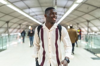 Young black man with backpack walking through bright modern airport walkway, smiling confidently as