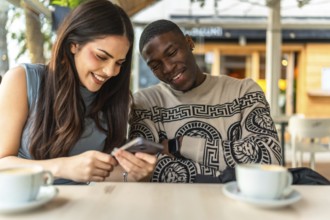 Diverse interracial couple smiling and sharing content on a smartphone at an outdoor cafe, enjoying