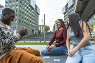 Diverse young friends sitting in a modern urban square, laughing and playing cards together