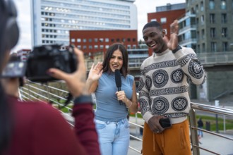 Journalist holding a microphone interviewing a young man on a city bridge, both waving and smiling