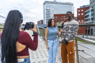 Woman filming an outdoor interview with a young woman holding a microphone to a laughing man,