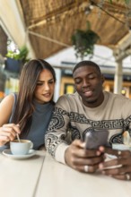 Diverse young woman and man sitting at an outdoor cafe table, smiling and sharing coffee while