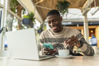 Young black man in a coffee shop using a laptop and smartphone, stirring his coffee while smiling,
