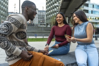 Diverse young friends laughing together while sitting and playing cards outdoors in a modern urban