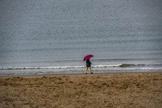 Person with red umbrella walking along the beach, calm sea in the background, winter break, on