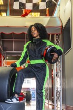 Young black woman standing confidently in a green and black race suit, holding a helmet and resting
