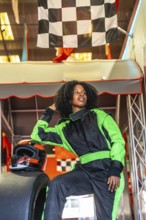 Confident young black woman in a green and black racing jumpsuit and helmet, posing near a kart