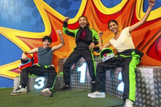 Three young multi ethnic friends on a karting podium, smiling and raising arms in victory while