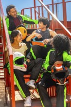 Happy diverse friends group wearing karting suits and helmets, sitting on metal stairs, laughing