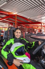 Young hispanic woman wearing a racing suit and holding a helmet, sitting in a go kart on the track,