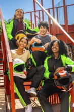 Diverse friends in racing suits holding helmets, smiling and sitting on red metallic stairs
