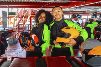 Multiracial young couple smiling at camera in racing suits and helmets, standing on indoor go kart
