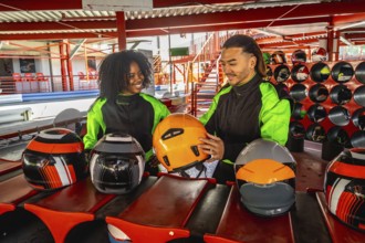 Diverse couple wearing racing suits and choosing helmets, standing on a go kart track, anticipating