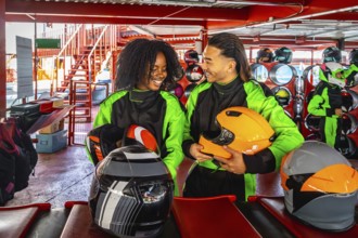 Happy young multiracial couple standing in an indoor karting track area, holding helmets, and