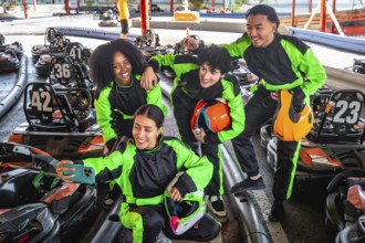 Diverse group of friends in racing suits and helmets smiling for a selfie at a karting track,