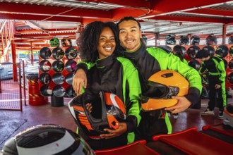 Young couple in racing suits with helmets, smiling at the camera on an indoor go kart track, ready