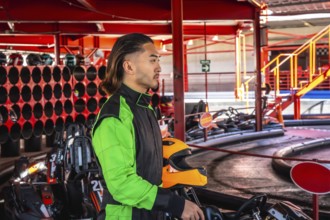 Young man wearing a racing suit standing with an orange helmet by a go kart, preparing to drive on