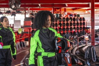Two cheerful young women in racing suits and holding helmets preparing to race go karts at an
