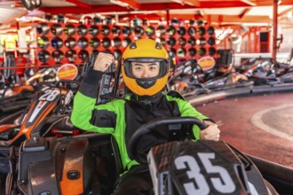 Asian man in racing suit and yellow helmet sits in a go kart on track, smiling and pumping fist in