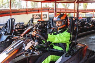 Young woman in a racing suit and helmet sitting in her go kart at the starting line, smiling and