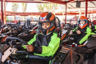 Young woman smiling in a black and neon green racing suit and orange helmet, sitting in a go kart