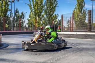 Drivers are speeding go karts around a curve on an outdoor asphalt track, wearing protective