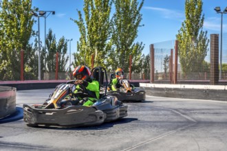 Two children in helmets and protective gear race go karts on an outdoor track, enjoying the speed,