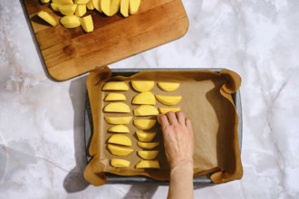 A hand is placing sliced potatoes on a baking tray lined with parchment paper. A wooden cutting