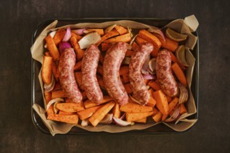 Raw sausages are placed on a baking tray with sliced sweet potatoes, onions, and garlic. The scene