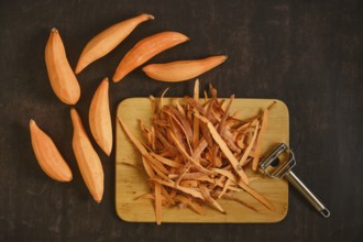 Peeled sweet potatoes with a vegetable peeler on a wooden cutting board. The skin is piled next to