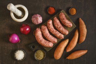 Natural casing sausages on a wooden board alongside sweet potatoes, garlic, onions, and various