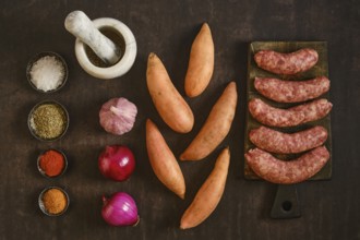 Natural casing sausages are placed next to sweet potatoes and several bowls of spices on a wooden