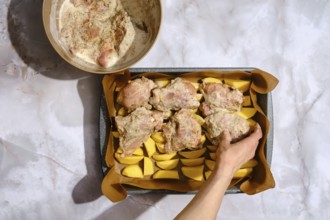A person prepares a dish by placing seasoned chicken on a tray lined with potatoes. There is a bowl