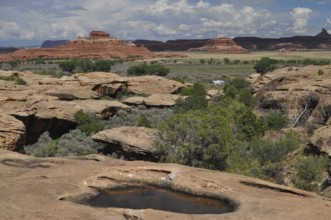 Wide rocky landscape with natural water basins and large red rocks in the background under a cloud