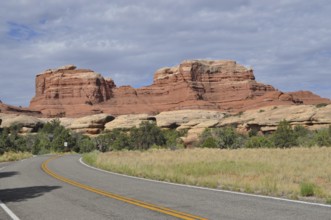 A winding country road runs in front of an impressive red rock formation under cloudy skies,