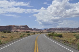 A road leads through vast desert landscape with impressive rock formations and cloudy skies,