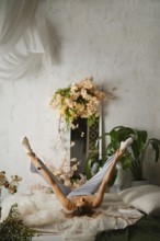 A woman is lying on a bed and stretching her legs in the air. The room has flowers and green plants
