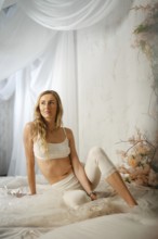 A woman in top and leggings sits on a bed and looks to her left. Soft light falls on her from above