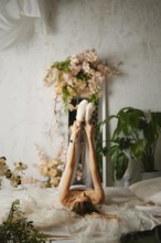 A woman is lying on her back while stretching her legs up in the air. The room has many plants and