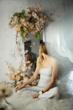 A woman sits cross-legged in a bright room. She faces a mirror decorated with flowers. Light