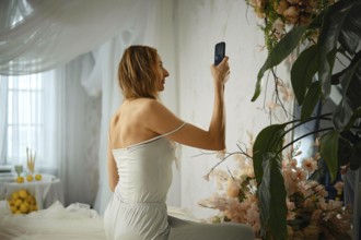 A woman sits in a room with light curtains and floral decorations, holding her phone to take a