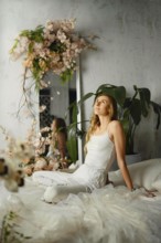 Dreamy woman with long hair sits on a bed in a light room. She looks up with a thoughtful