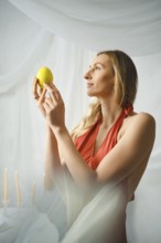 A woman stands indoors, holding a lemon in her hands. She gazes at it with focus. Soft light