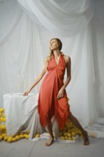 A woman stands near a table covered with a white cloth. She poses in a simple dress. Around her are