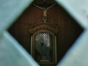 Wooden statue of Jesus on the cross through a grid in a dark chapel, hiking in the Frankenwald