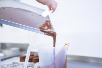 Liquid chocolate is poured from a tray into a large plastic container, presumably in a kitchen,