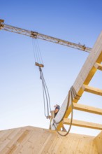 Worker handles a rope next to a crane under a blue sky on a construction site, timber house